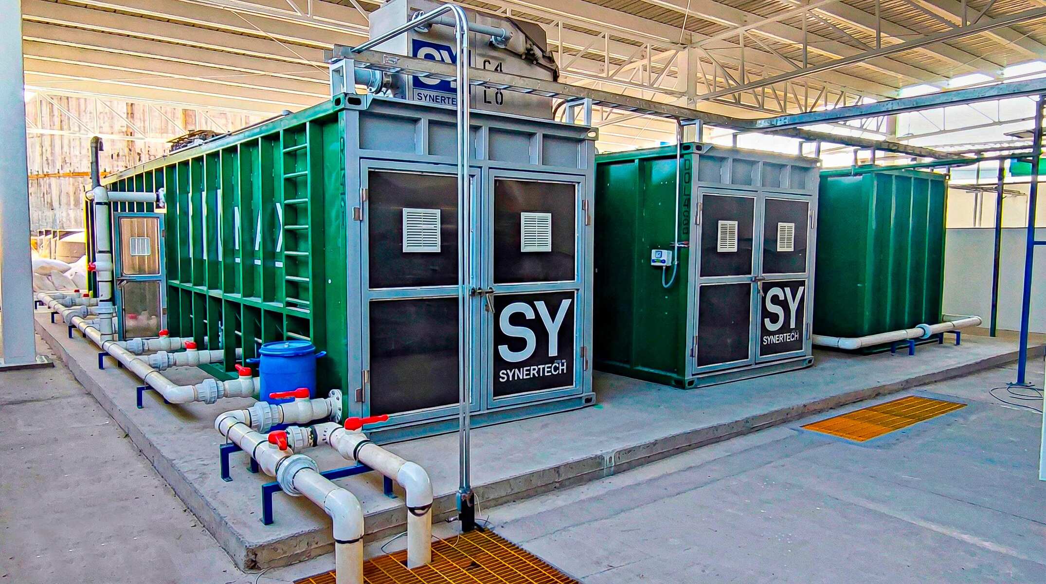 Técnico inspeccionando una planta de tratamiento de aguas residuales modular de color verde marca BIOMAX SYNERTECH, que incluye módulos de pretratamiento, decantación, DAF y oxidación avanzada. La planta está ubicada en un entorno industrial con techo alto y tuberías visibles. - ver más información